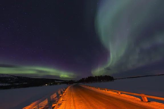 Grüne Polarlichter über einer verschneiten Straße und zugefrorenem See bei Arjeplog in Nordschweden während der Wintererprobung.