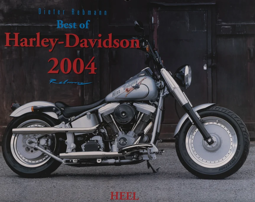 Harley-Davidson Softail Fat Boy mit „Miss Behavin'“-Tankdesign in Seitenansicht vor dunkler Industriehalle.Harley Kalender 2004