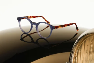 Runde Designerbrille mit blauem Rahmen und gemusterten Bügeln auf der Motorhaube eines Oldtimers neben einem Scheinwerfer.