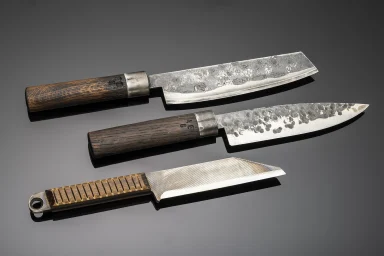 Drei handgeschmiedete Messer mit Holzgriffen und unterschiedlichen Stahlklingen auf grauem Hintergrund.