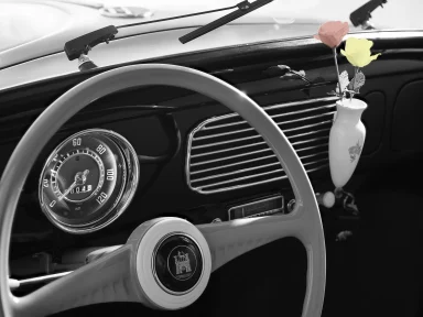 Innenraum eines klassischen Volkswagen Käfers mit weißem Lenkrad, geöffneten Türen und schlichtem Armaturenbrett in Schwarz-Weiß-Darstellung